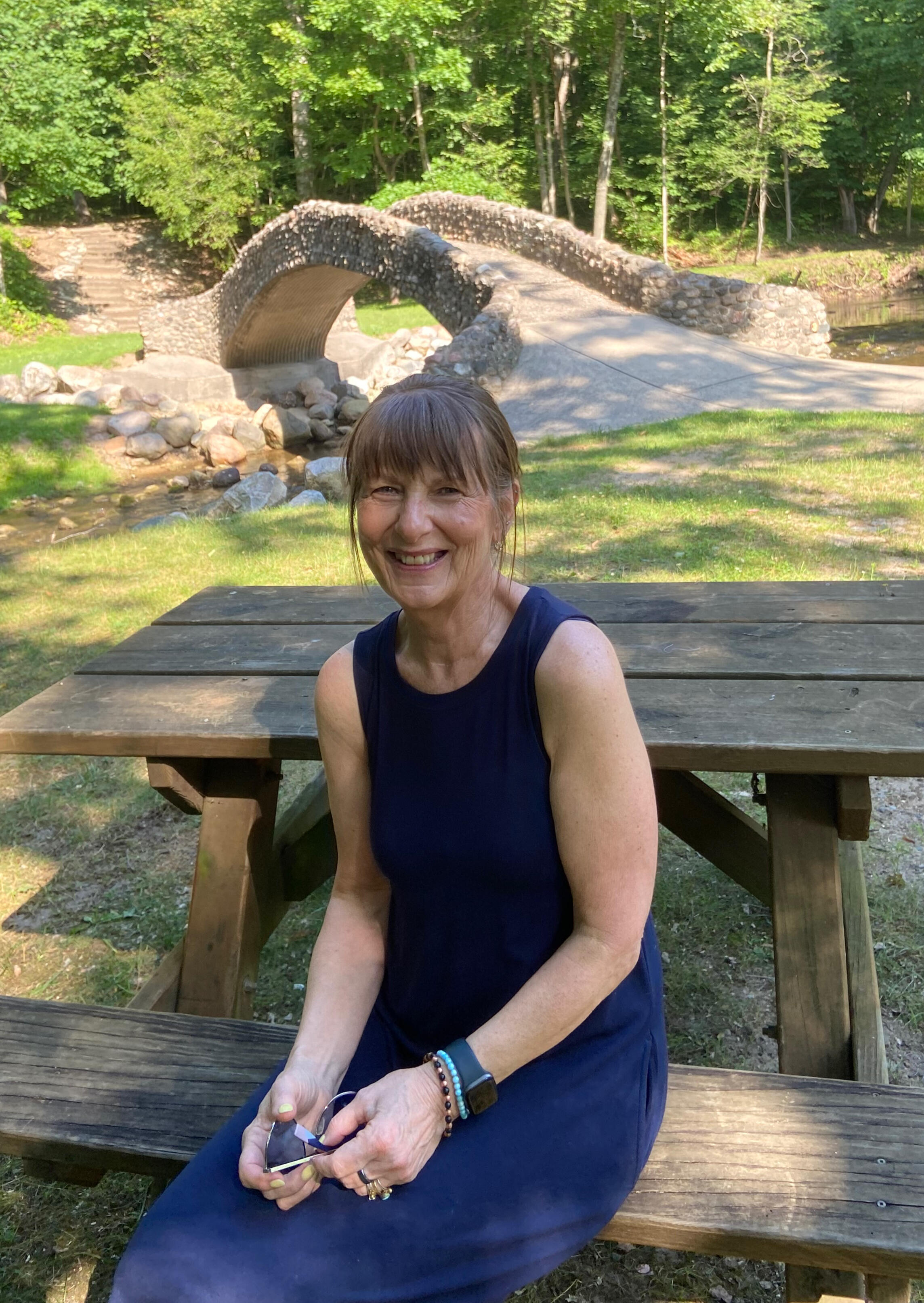 Terry Pershon sitting at picnic table in the park with stone bridge behind her in a serene peaceful setting